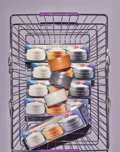 A wire shopping basket containing boxes and stacked jars of Fenty Skin skincare products, set against a pale purple background.