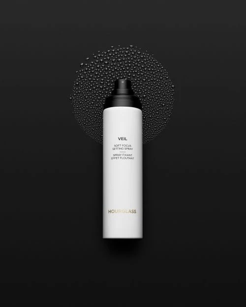 Hourglass Veil Soft Focus Setting Spray in a white bottle with black top, against a black background with water droplets.
