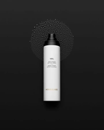 Hourglass Veil Soft Focus Setting Spray in a white bottle with black top, against a black background with water droplets.