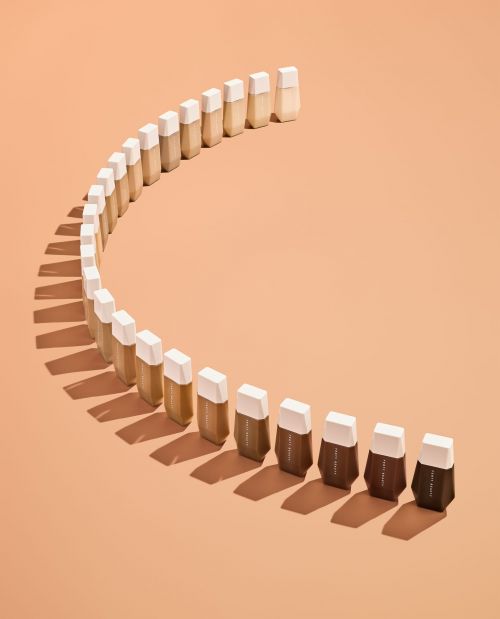 Makeup foundation bottles arranged in a curve, showcasing a gradient of shades from light to dark on a peach-colored background.