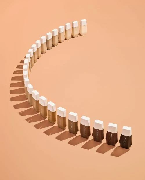 Makeup foundation bottles arranged in a curve, showcasing a gradient of shades from light to dark on a peach-colored background.