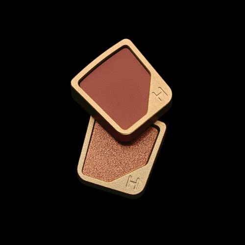 Two makeup compacts stacked on a black background in a product shot showcasing a gold rimmed, square case with a brown and bronze fill.