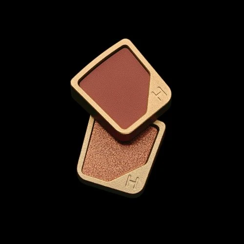 Two makeup compacts stacked on a black background in a product shot showcasing a gold rimmed, square case with a brown and bronze fill.