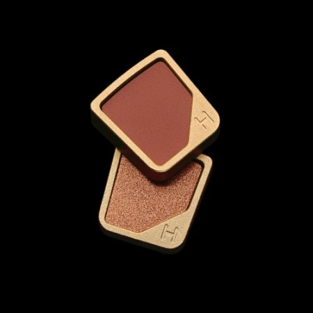 Two makeup compacts stacked on a black background in a product shot showcasing a gold rimmed, square case with a brown and bronze fill.