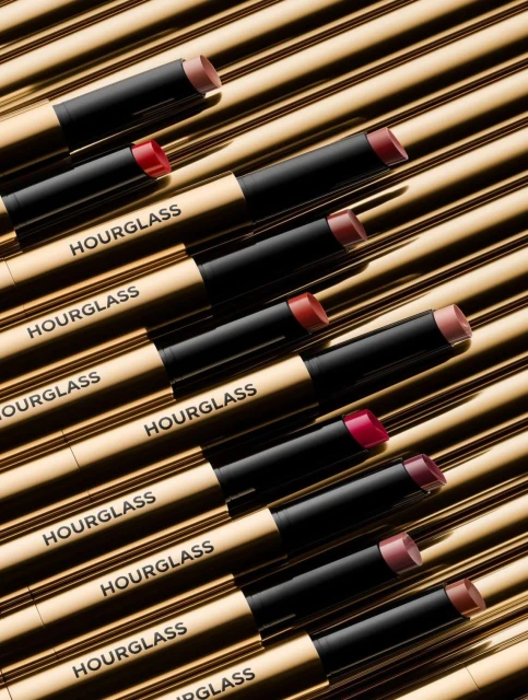 Close-up of multiple gold-toned Hourglass lipsticks with black caps, arranged diagonally against a striped gold background. Beauty product shot.