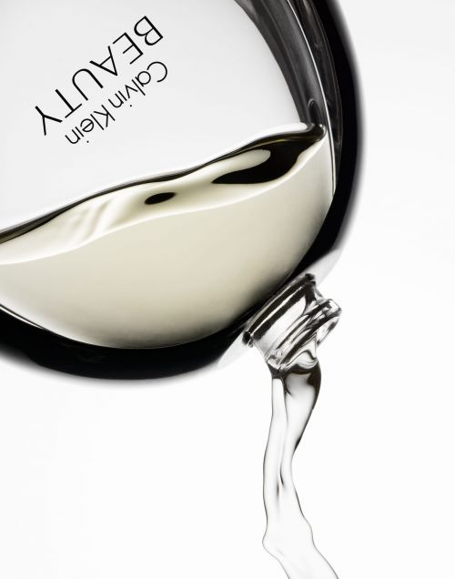Close-up of a Calvin Klein Beauty perfume bottle tilted to pour liquid, set against a plain white background in a beauty advertisement.