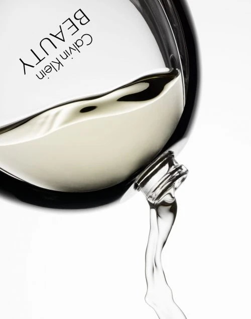 Close-up of a Calvin Klein Beauty perfume bottle tilted to pour liquid, set against a plain white background in a beauty advertisement.