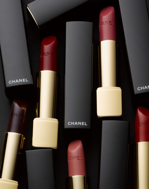 Several Chanel lipsticks in black cases are displayed on a black background, featuring red and rose-colored lipstick shades.