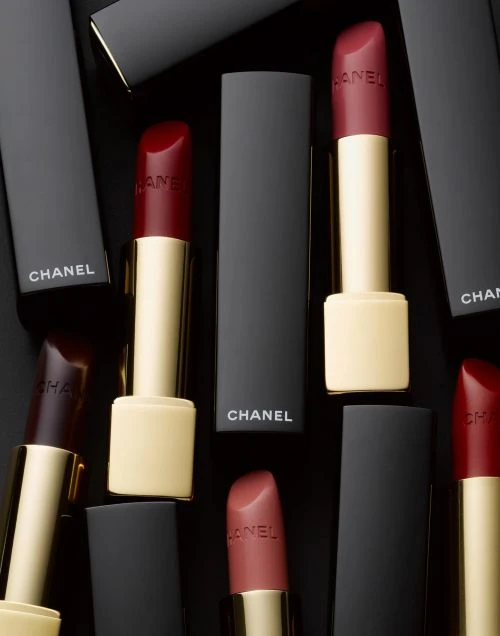 Several Chanel lipsticks in black cases are displayed on a black background, featuring red and rose-colored lipstick shades.