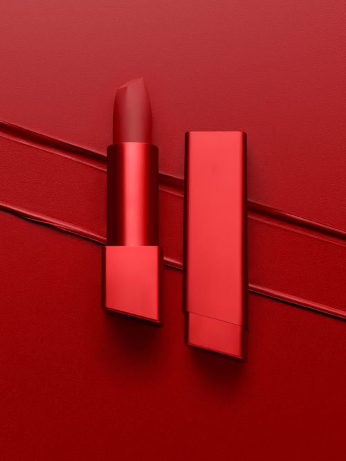 Studio shot of a bold red lipstick with its cap off sitting on a matching red textured surface with lipstick smear details.