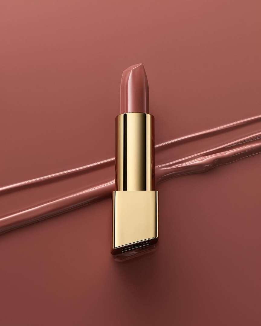 Close-up studio shot of a single lipstick with a gold tube on a tan backdrop with strokes of lipstick, beauty and makeup concept.