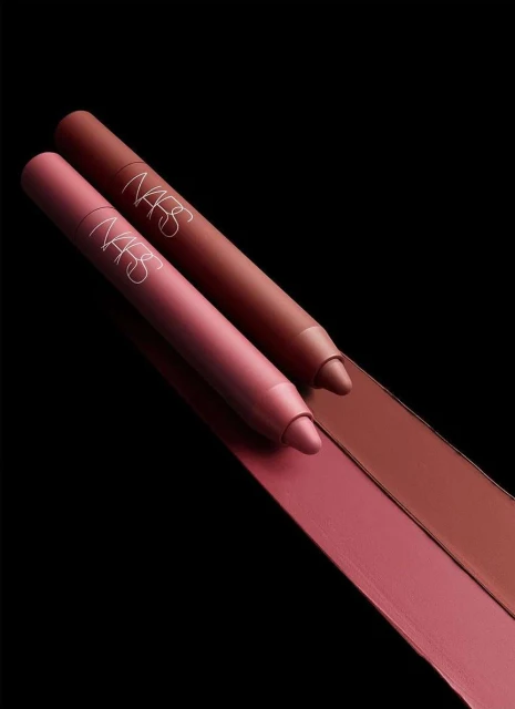 Close-up of two NARS lip crayons in pink and brown hues with matching lipstick swatches on a dark background, beauty product photography.