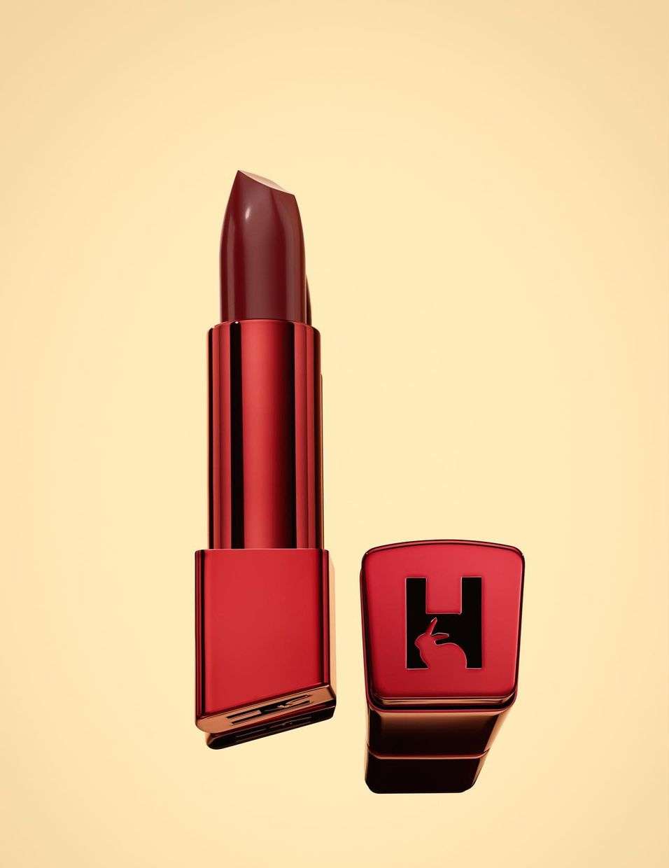 Close-up of a red lipstick tube and its cap, featuring a logo with a rabbit and the letter H, on a light tan background.