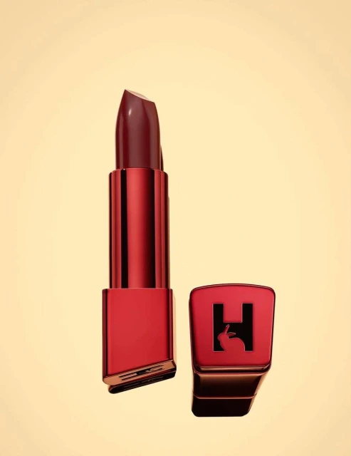 Close-up of a red lipstick tube and its cap, featuring a logo with a rabbit and the letter H, on a light tan background.