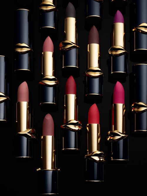 An array of Pat McGrath Labs lipstick tubes in various colors, accented with gold lips, arranged on a sleek black backdrop.