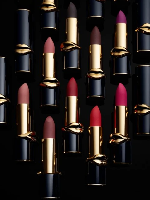 An array of Pat McGrath Labs lipstick tubes in various colors, accented with gold lips, arranged on a sleek black backdrop.