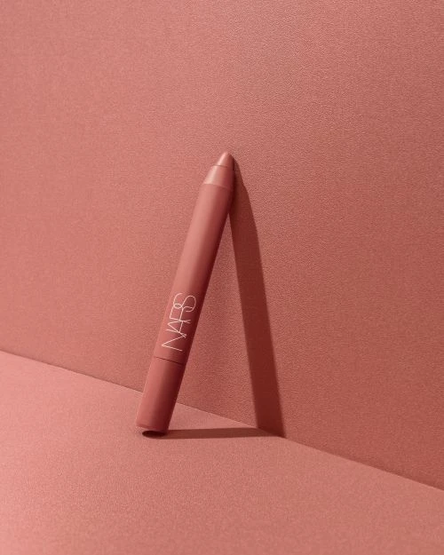NARS lipstick crayon standing against a monochromatic coral-pink background in a beauty product shot. Makeup and cosmetic photography.