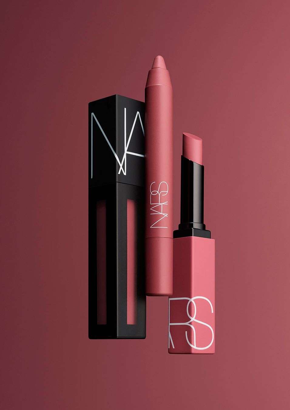 Three NARS cosmetics products: lipstick, lip crayon, and liquid lip, on a solid rose-colored background in a studio shot.