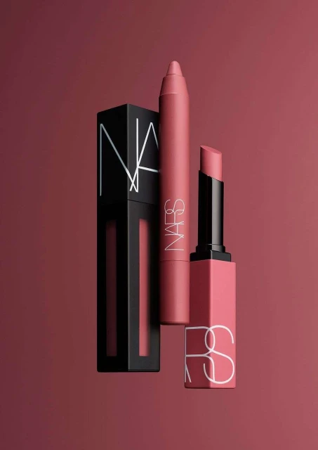 Three NARS cosmetics products: lipstick, lip crayon, and liquid lip, on a solid rose-colored background in a studio shot.