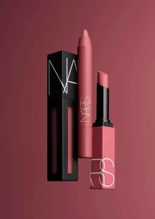 Three NARS cosmetics products: lipstick, lip crayon, and liquid lip, on a solid rose-colored background in a studio shot.