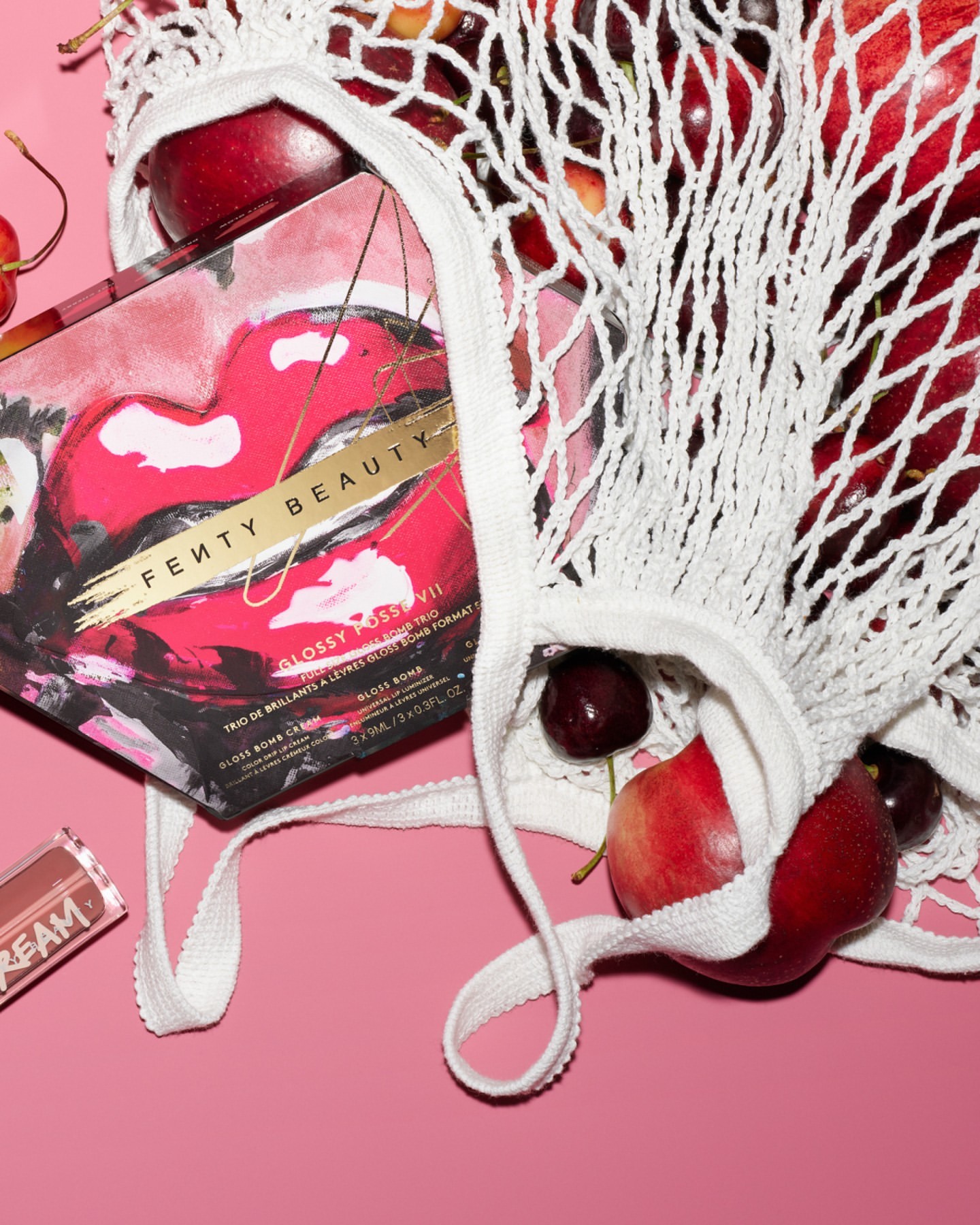 Fenty Beauty makeup box inside a white mesh bag filled with red cherries and apples on a pink background, flat lay arrangement.