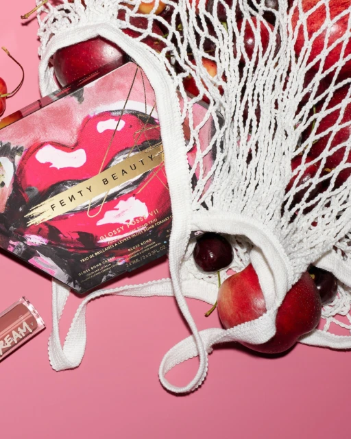 Fenty Beauty makeup box inside a white mesh bag filled with red cherries and apples on a pink background, flat lay arrangement.