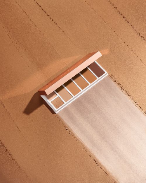 Makeup palette of neutral and brown eyeshadows displayed against a textured tan background, showcasing beauty product packaging.