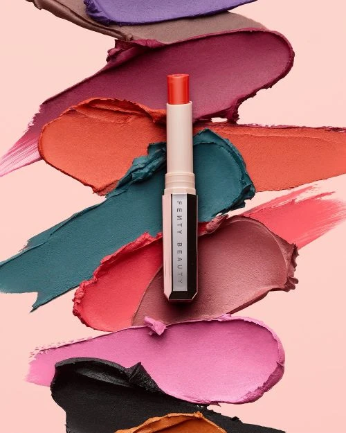 Product shot of coral lipstick tube surrounded by swatches of vibrant makeup in pink, purple, coral, green, brown against light pink.