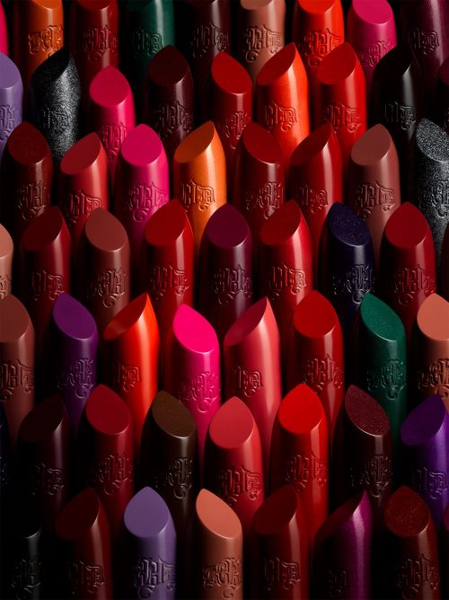 A close-up studio shot features an array of Kat Von D lipsticks in various shades including red, pink, purple, brown, and green, arranged in a tight composition.