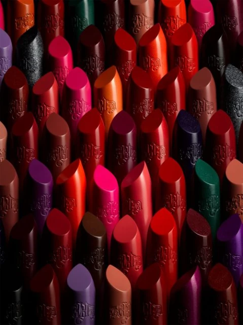 A close-up studio shot features an array of Kat Von D lipsticks in various shades including red, pink, purple, brown, and green, arranged in a tight composition.
