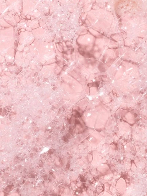 Close-up of translucent, iridescent soap bubbles of varying sizes against a soft pink background creating a textured, ethereal effect.