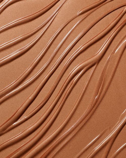 Close-up of textured, wavy smears of bronzer makeup or skincare product. The surface is glossy and reflective, with soft, warm tones.