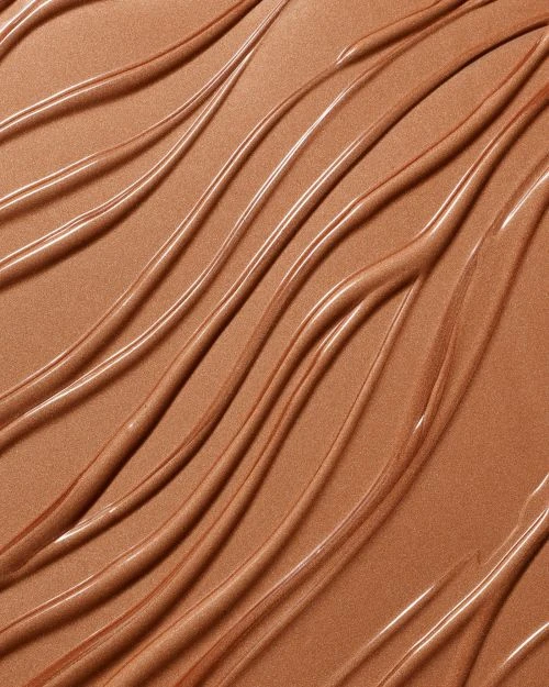Close-up of textured, wavy smears of bronzer makeup or skincare product. The surface is glossy and reflective, with soft, warm tones.