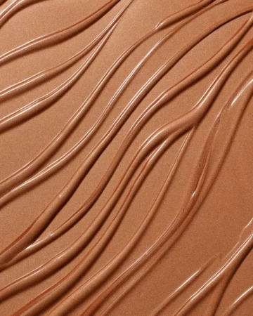 Close-up of textured, wavy smears of bronzer makeup or skincare product. The surface is glossy and reflective, with soft, warm tones.