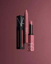 Product photograph of NARS cosmetics including a black box, a matte lip crayon, and a lipstick against a mauve background.