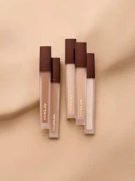 Five upright liquid concealer tubes with dark brown caps arranged vertically on a soft beige fabric background.