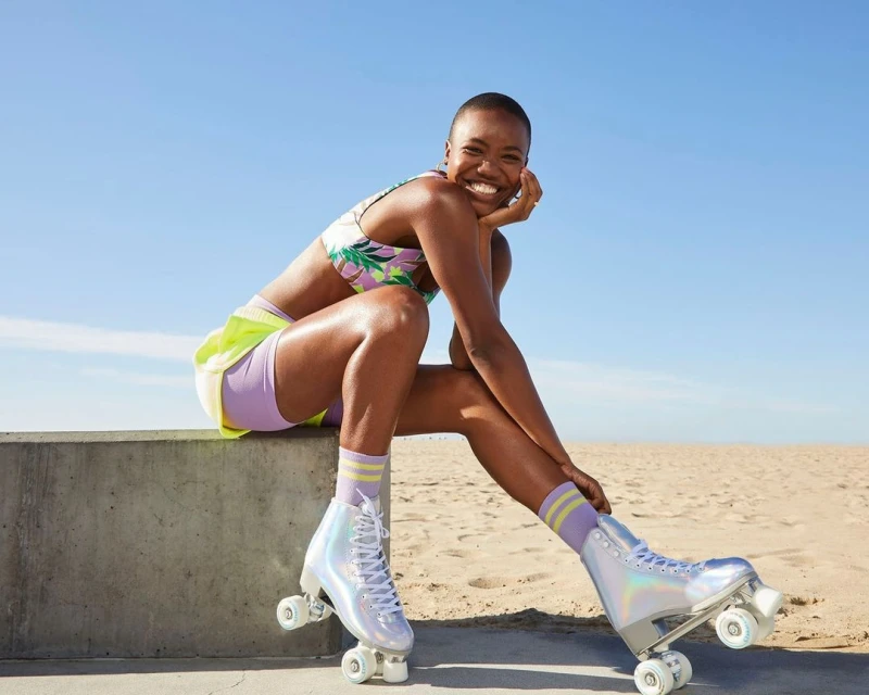 Athletic African American woman with roller skates smiles sitting outside in the sunlight with blue sky and beach in background.