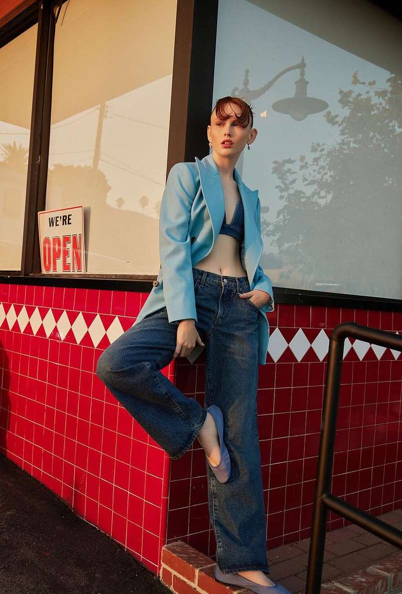 A young adult woman in a blue blazer, denim jeans, and blue shoes, leans against a red-and-white tiled wall outside a building with a sign.