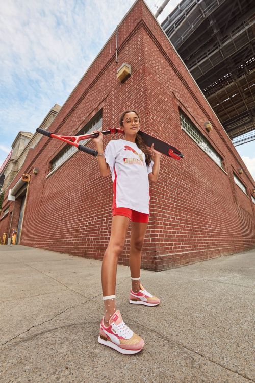 A young person stands in an urban setting, wearing Puma athletic apparel, holding a scooter over their shoulders, and wearing sneakers.