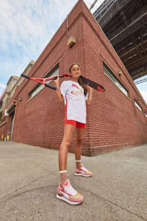 A young person stands in an urban setting, wearing Puma athletic apparel, holding a scooter over their shoulders, and wearing sneakers.