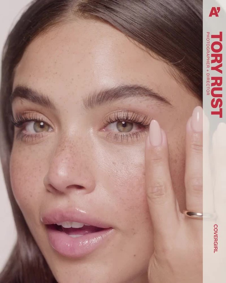 Close-up of a light-skinned adult female with freckles and neutral expression, wearing a gold ring, applying makeup near her eye.