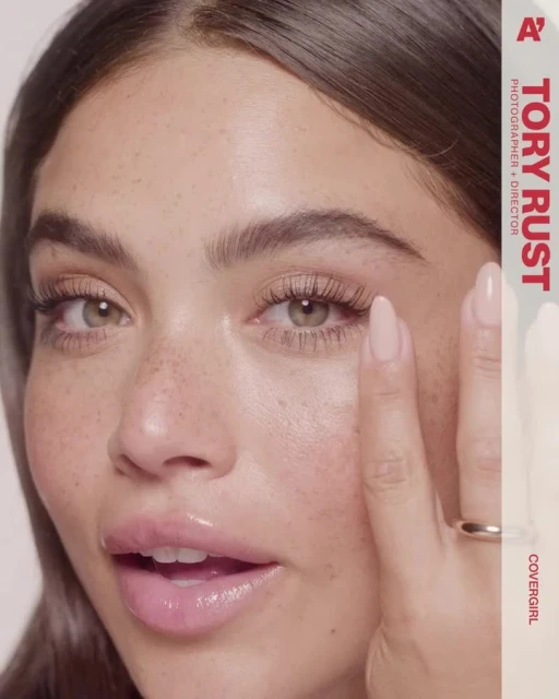 Close-up of a light-skinned adult female with freckles and neutral expression, wearing a gold ring, applying makeup near her eye.