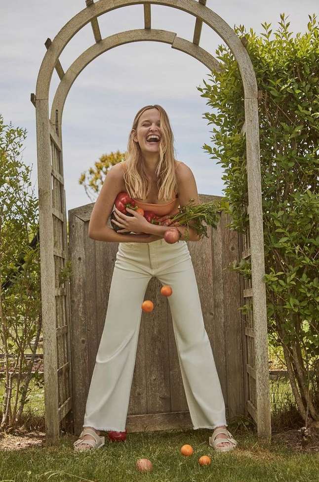 Smiling blonde woman holding fresh produce and juggling small citrus fruits under a wooden garden arch by Tory Rust.