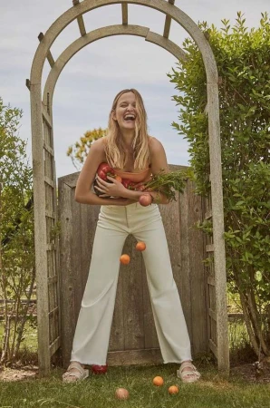 Smiling blonde woman holding fresh produce and juggling small citrus fruits under a wooden garden arch by Tory Rust.