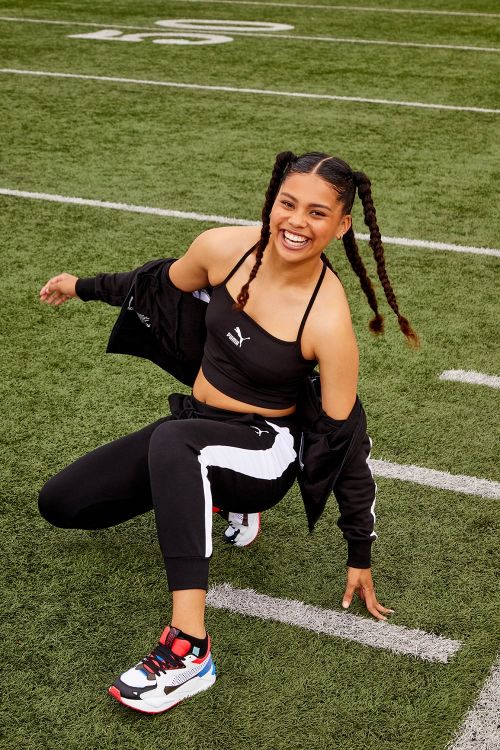 A smiling woman in black Puma workout gear poses on a green football field with white yard lines, exuding confidence and athleticism.