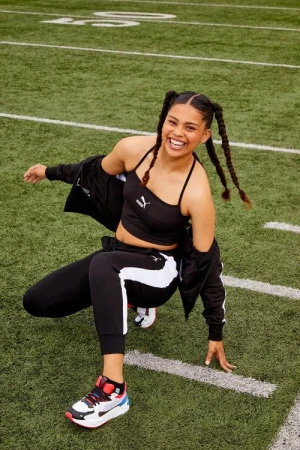 A smiling woman in black Puma workout gear poses on a green football field with white yard lines, exuding confidence and athleticism.