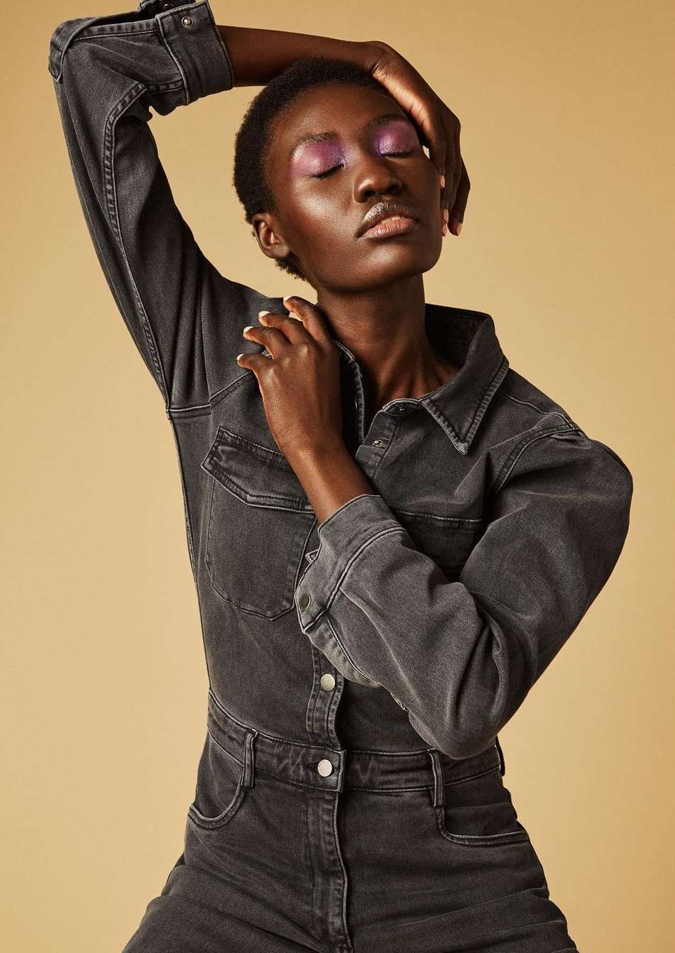 An adult female model with dark skin poses in a grey denim jumpsuit against a neutral background, showcasing beauty and fashion in studio lighting.