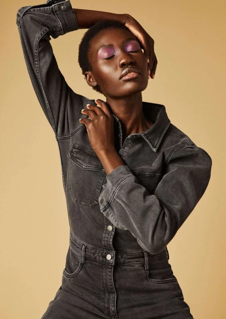 An adult female model with dark skin poses in a grey denim jumpsuit against a neutral background, showcasing beauty and fashion in studio lighting.