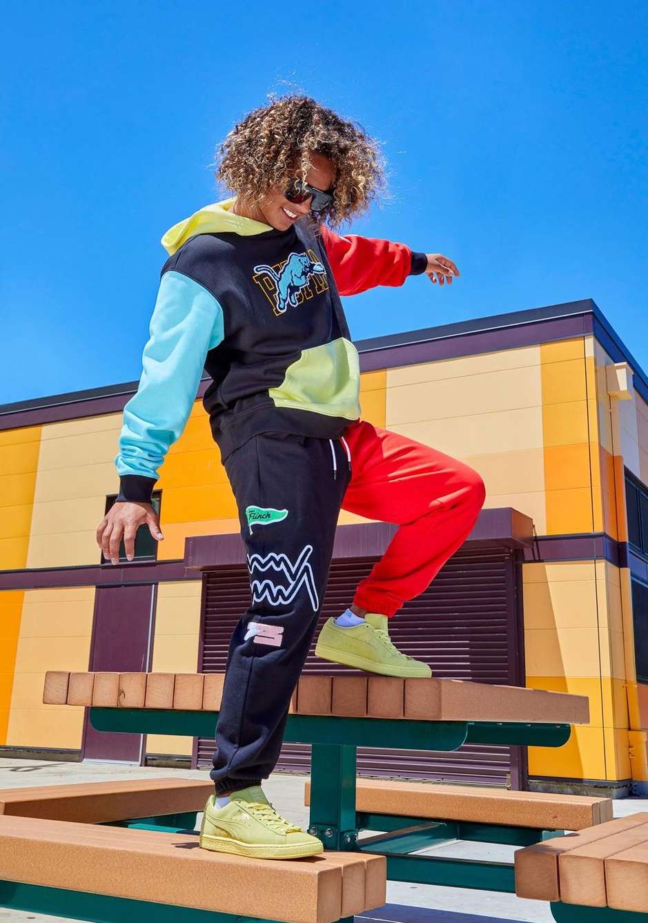 A young adult stands on a picnic table in colorful sportswear against a bright blue sky and a modern orange and brown building.