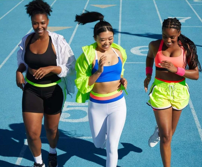 Three smiling women wearing athletic clothing jog on a bright blue running track in the sunshine, promoting fitness and healthy lifestyle.